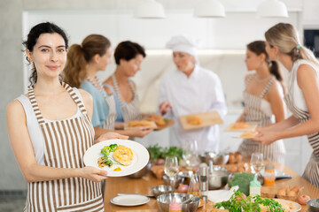 Young participiant of cooking master class holding plate with cooked chicken breast in her hands standing around other female members