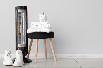 Electric heater and piggy bank with clothes on stool near white wall. Heating season concept