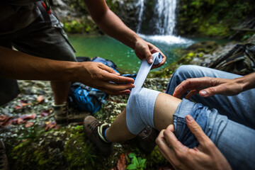 Assistance of a Woman Thigh With an Elastic Medical Bandage from First Aid Travel Kit on a Hike in Wilderness - obrazy, fototapety, plakaty