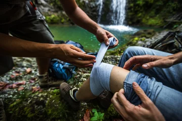 Fotobehang Assistance of a Woman Thigh With an Elastic Medical Bandage from First Aid Travel Kit on a Hike in Wilderness © Fotopogledi