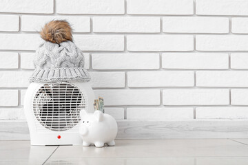 Electric fan heater with piggy bank and hat near white brick wall. Heating saving concept
