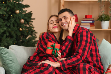 Young couple wearing plaid pajamas with gift in living room decorated for Christmas