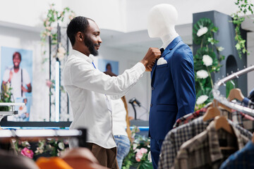 Clothing store worker fixing tie on mannequin wearing formal suit, showcasing menswear to...