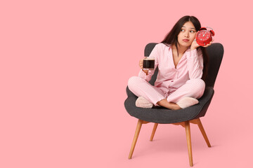 Young Asian thoughtful woman in pajamas with alarm clock and cup of coffee sitting on armchair against pink background