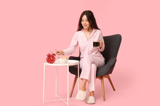 Young Asian woman in pajamas with alarm clock, cup of coffee and cornflakes sitting on armchair against pink background