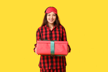 Beautiful young Asian woman in checkered pajamas with sleeping mask and Christmas gift box on yellow background