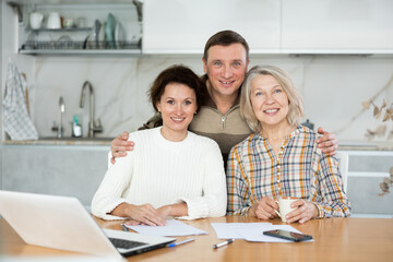 Obraz premium Smiling middle-aged man and two women posing at the kitchen-table with sheets of paper and laptop in front of them