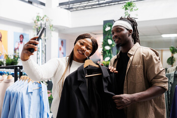 Man and woman influencers taking smartphone selfie to promote new line of clothing in shopping mall. Online bloggers holding jacket on hanger and making photo for followers in fashion boutique