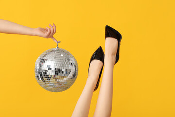Female legs in trendy high heels and hand with disco ball on yellow background
