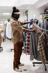 Foto auf Alu-Dibond African american man browsing clothes in retail store showroom. Young customer checking apparel hanging on rack to buy casual plaid shirt in shopping mall fashion boutique © DC Studio