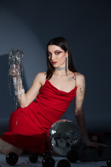 Young woman with disco ball and glass of Christmas tinsel sitting on grey background