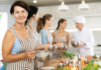 Aged female participiant of cooking master class holding bowl and whisk in her hands standing around other female members