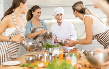 Elderly woman cook in white suit holds bowl in hands and tells women students of culinary courses...