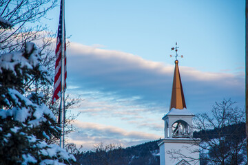 North Conway NH White church belfry with copper steeple and weathervane against blue sky with American flag and snow-covered trees in foreground