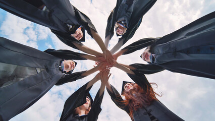 Group of happy successful graduates in academic hats and robes standing in circle and putting their...