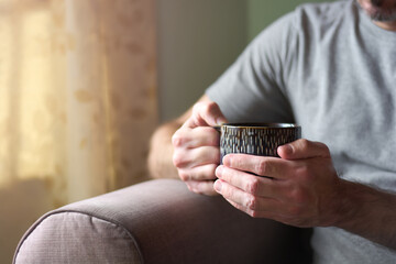 Close up of hands of young caucasian man holding a cup of tea. Enjoying serene morning with hot tea in his living room at home, relaxing during breakfast. Copy space for text