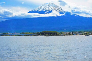 Fujiyama mountain before the lake with trees, houses, buildings, bridge, and ships under cloudy-blue sky background 