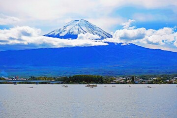 Fujiyama mountain before the lake with trees, houses, buildings, bridge, and ships under cloudy-blue sky background 