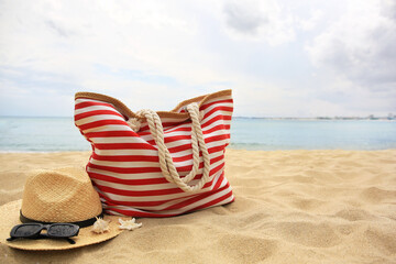 Stylish striped bag with straw hat, sunglasses, seashell and starfish on sandy beach near sea....