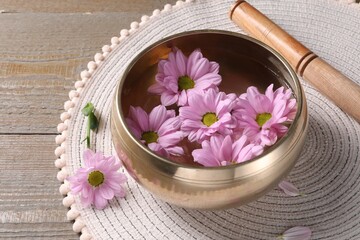Tibetan singing bowl with water, beautiful flowers and mallet on wooden table, closeup