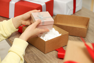 Woman putting Christmas gift box into parcel at wooden table, closeup. Sending present by mail