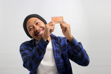 A happy young Asian man, dressed in a beanie hat and casual shirt, enthusiastically shows a credit...