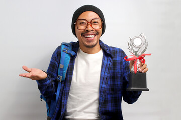 Happy young Asian student, adorned in a beanie hat, eyeglasses, and a casual shirt, proudly lifts his trophies while smiling and looking directly at the camera, while standing against white background
