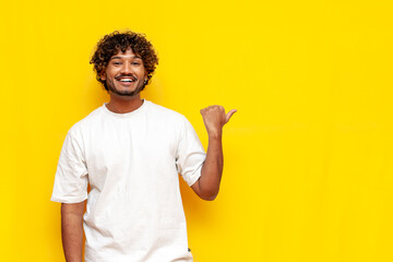 young indian curly guy pointing at copy space on yellow isolated background and smiling - obrazy, fototapety, plakaty