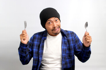 Hungry Asian man, dressed in a beanie hat and casual shirt, bites his lips while holding a fork and...