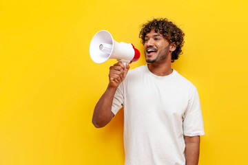 young indian guy announces information into a megaphone on a yellow isolated background - obrazy, fototapety, plakaty