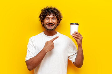 young indian guy pointing at paper cup of coffee on yellow isolated background, south asian man in...
