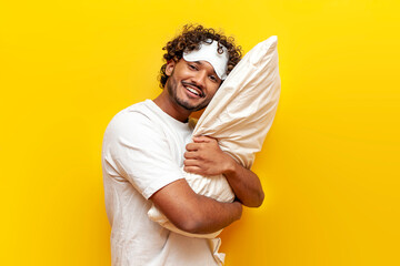 young indian sleepy guy in pajamas and eye mask hugging soft pillow on yellow isolated background