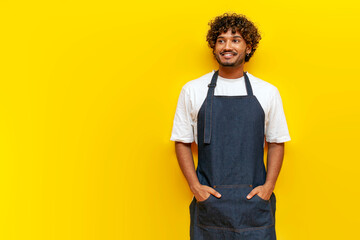 young indian guy barista in an apron stands and looking at copy space on yellow isolated background