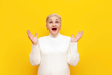 shocked old granny in a white sweater wonders with her mouth open over yellow isolated background