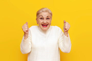 cheerful old grandmother in white sweater rejoices in victory and luck on yellow isolated background