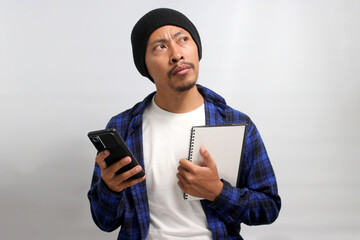 Pensive young Asian man, dressed in a beanie hat and casual shirt, holds a mobile phone and a notebook, gazing toward the copy space, deep in thought, seemingly seeking inspiration or ideas