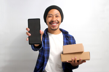 Excited young Asian man, dressed in a beanie hat and casual clothes, showcases a smartphone with an...