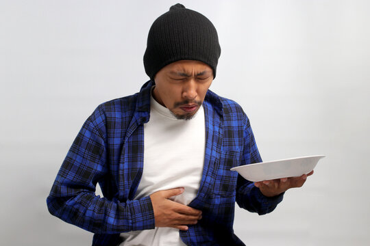 Distressed young Asian man, clutching his stomach and an empty white plate, expressing nausea and stomach pain likely caused by an allergic reaction, food poisoning, or consuming unhealthy food