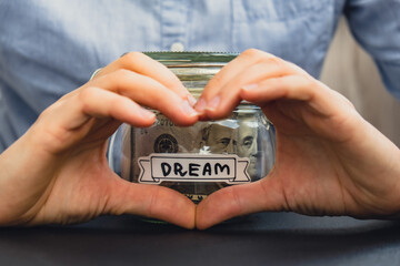 Unrecognizable woman showing heart sign Saving Money In Glass Jar filled with Dollars banknotes. DREAM transcription in front of jar. Managing personal finances extra income for future insecurity