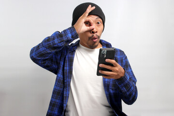 A young Asian man, dressed in a beanie hat and casual clothes, makes an OK sign gesture while holding his smartphone, conveying agreement, approval, and satisfaction, indicating a positive review