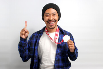Asian man, dressed in a beanie hat and casual shirt, points his finger upward with an expression...