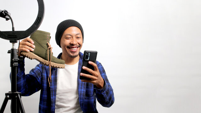 A young Asian influencer, dressed in a beanie hat and casual shirt, unboxes shoes, shares a review, and captures content for social media while standing against a white background