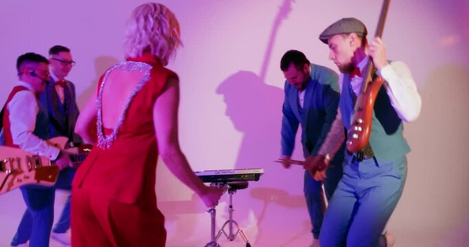 Musc band playing instruments dancing performing on stage having fun together. Blonde woman singer in red costume with microphone. Men musicians with musical instruments. Performance, show concept.