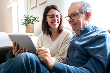 Happy senior man and his young daughter laughing together using digital tablet sitting on sofa at...