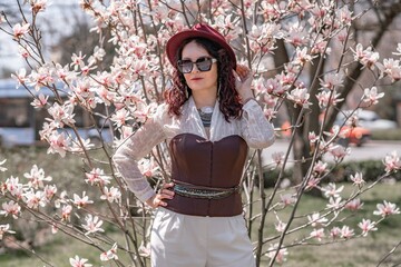 Magnolia park woman. Stylish woman in a hat stands near the magnolia bush in the park. Dressed in...