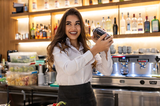Young female worker at bartender desk in restaurant bar preparing cocktails with shaker. Beautiful young woman behind bar