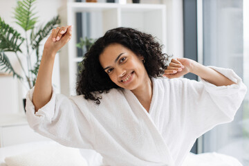 Young female adult in white dressing gown stretching her limbs while resting on cozy bed linen in...