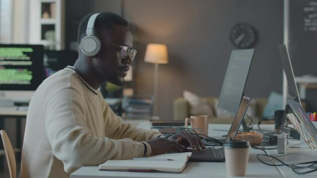 Side view waist up shot of young African American male programmer wearing wireless headphones and eyeglasses working on laptop in minimalist officeSide view waist up shot of young African American mal