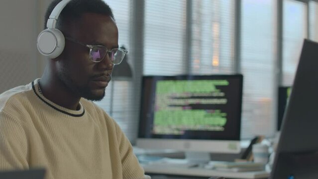 Waist up of young African American male software engineer wearing wireless headphones and eyeglasses working on desktop computer in modern office