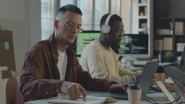 Waist up of two young multiracial software engineers working on laptop and computer while sitting at one table in minimalist programmers office